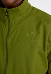 Close-up of green jacket with zip and embroidered The North Face logo on upper chest against a white background.