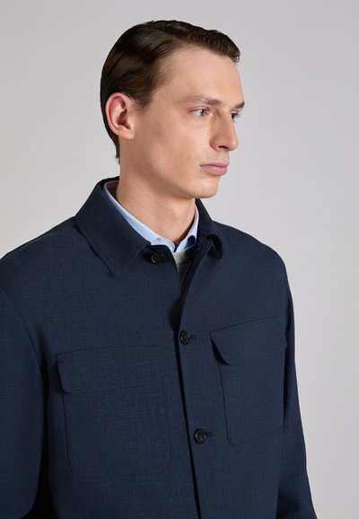Młody mężczyzna z zaczesanymi do tyłu włosami, ubrany w granatową, zapinaną na guziki kurtkę na jasnoniebieską koszulę z kołnierzykiem, spoglądający na bok na tle szarego tła.