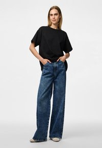 Svart oversize T-shirt med korta ärmar kombinerad med vida, högmidjade blå denimjeans. Modellen står med händerna i fickorna.
