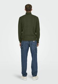Jersey verde trenzado con cuello alto, combinado con jeans cargo azules con bolsillos laterales y un corte holgado. Zapatillas blancas completan el look.