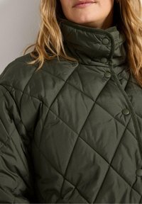 Una donna indossa un giubbotto acolcilato verde con un alto colletto e bottoni, mostrando parte del suo viso e i lunghi capelli ondulati.