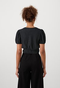 Top nero con texture caratterizzato da maniche corte a palloncino e vita smock, visto da dietro. Abbinato a pantaloni neri.