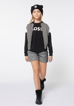 Veste bomber noire avec des manches à motifs, t-shirt noir avec le logo "BOSS", short gris et bottes noires à lacets. Bonnet en tricot noir sur la tête.