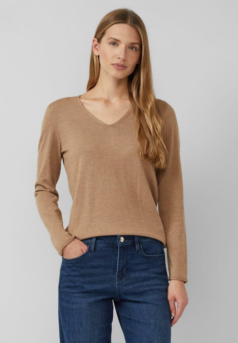 Femme aux longs cheveux blonds portant un pull en V couleur camel et un jean bleu, se tenant avec une main dans la poche contre un fond gris.