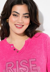 T-shirt di cotone rosa con scollo a V, caratterizzato da un orloconsumato e borchie metallizzate che formano la parola "RISE" sul davanti. Due bottoni sul collo.
