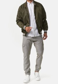 Veste aviateur vert olive avec poignets rayés blancs, associée à un pantalon cargo gris clair et des baskets blanches. Chemise blanche à col portée en dessous.