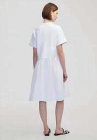 Witte katoenen jurk met korte mouwen, een ronde hals en een geplooide taille. De rok loopt soepel uit en reikt tot halverwege het kalf. Platte sandalen.