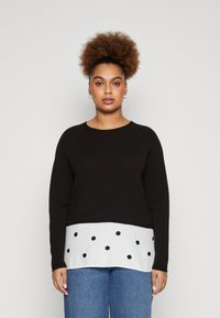 Maglione lungo nero a maniche lunghe con orlo bianco a pois. Presenta un collo a giro e maniche raglan. Indossato con jeans blu.