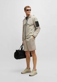 Beige lightweight jacket with zip pockets and hood, paired with matching shorts. Black duffel bag and neutral sneakers with accents.
