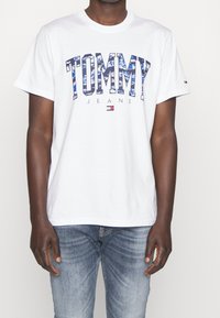 Bílý tričko s modrým logem "TOMMY" a vzorovaným designem, s nápisem "JEANS" pod ním. Krátké rukávy a klasický kulatý výstřih.