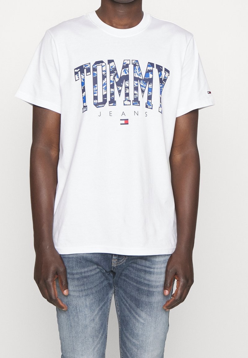 Bílý tričko s modrým logem "TOMMY" a vzorovaným designem, s nápisem "JEANS" pod ním. Krátké rukávy a klasický kulatý výstřih.