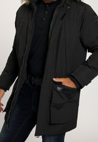 Veste noire matelassée avec une fermeture éclair à l'avant, capuche ajustable ornée d'une bordure en fausse fourrure, et poches latérales. Tissu texturé avec une finition lisse.