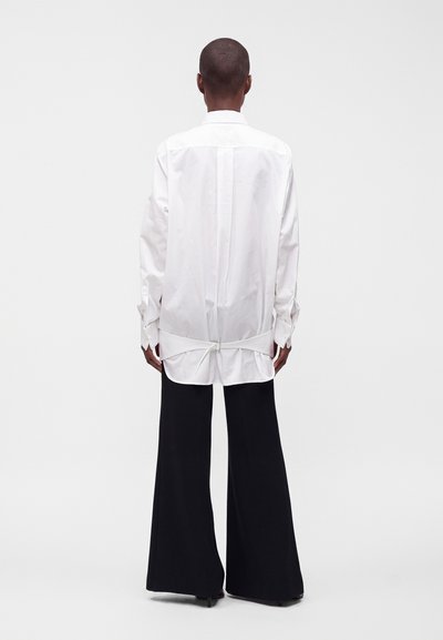 Personne vue de dos portant une longue chemise blanche avec un détail noué dans le dos et un pantalon noir à jambes larges sur un fond clair uni.
