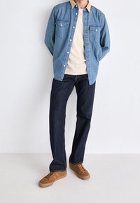 Camicia di jeans con colletto beige, due tasche sul petto, indossata sopra una camicia crema. I jeans blu scuro e le sneakers in suede marroni completano l'outfit.
