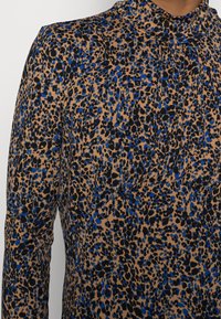 Blusa a maniche lunghe con uno sfondo beige decorato da macchie di Leopardo nere e blu, tessuto morbido e scollatura alta con delicate pieghe.