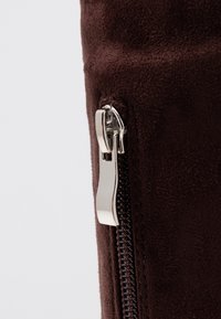 Hnědá semišová taška na zip má stříbrný kovový zip a texturovaný povrch, s jemně prošívaným obrysem a hladkým táhlem zipu.