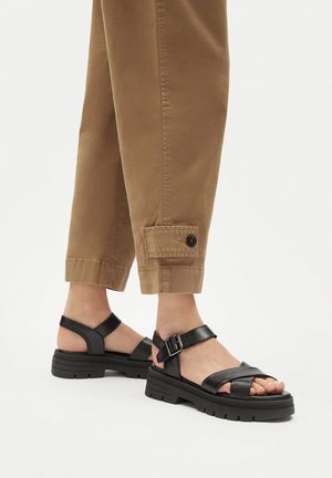 Schwarze Ledersandalen mit gekreuzten Riemen, verstellbarer Schnalle am Knöchel und dicker profilierter Sohle. Kombiniert mit hochgekrempelten braunen Hosen.