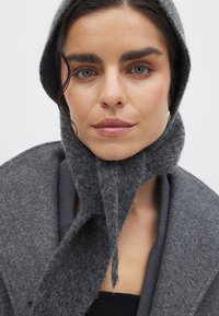 Hallhallane villasegu kootud mantel koos kinnitatud salliga, millel on tekstuurne viimistlus ja kapuuts. Kujundusel on struktureeritud krae ja nurkne kuju.