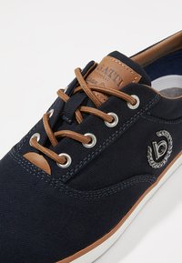 Navy canvas shoe with brown leather accents, round toe, metal eyelets, and contrasting white rubber sole. Features emblem on the side.