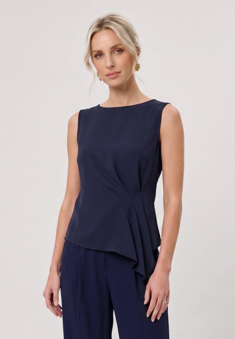 Femme blonde à la peau claire portant des boucles d'oreilles en or et un haut sans manches bleu marine avec des détails en couches, assorti à un pantalon bleu marine.
