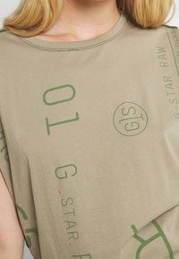 Close-up of a person wearing a beige t-shirt with green printed text and logos, including "G-STAR RAW" and a circular "GS" symbol.
