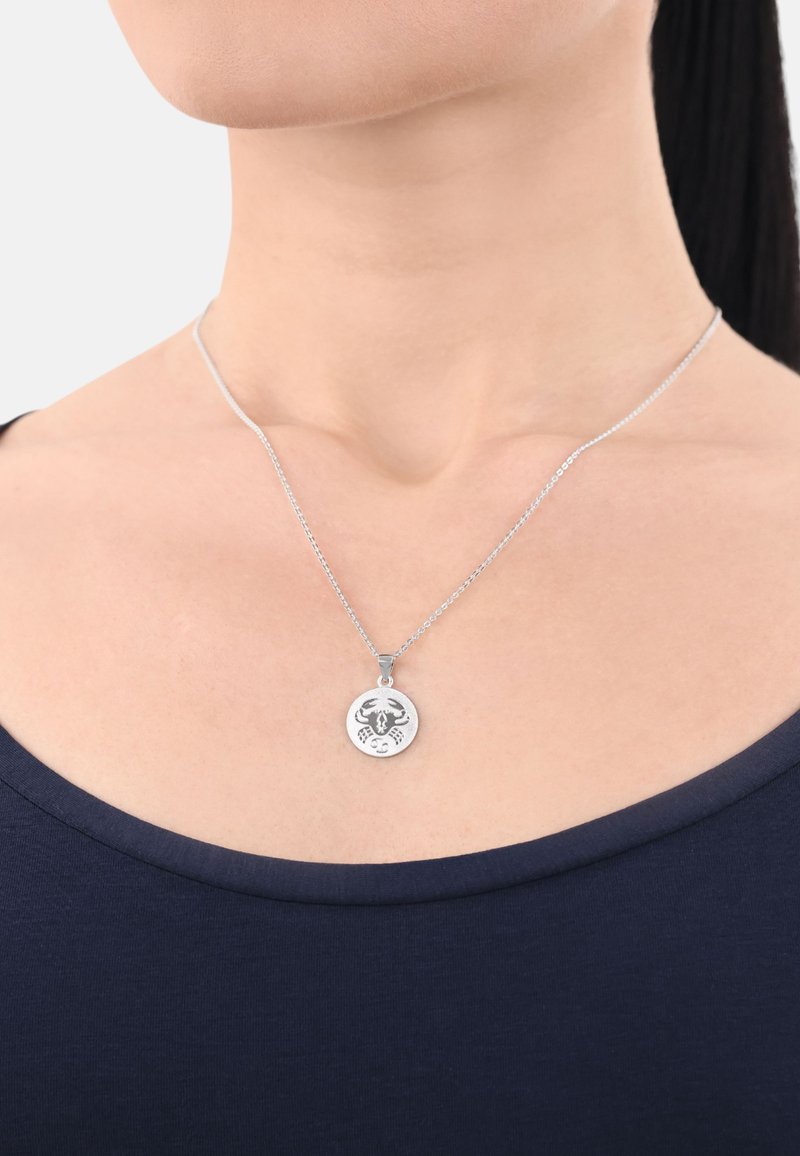 Silverhänge i form av en cirkel med en ingraverad skorpionmotif, hängande i en delikat kedja, som bärs över en mörk topp.