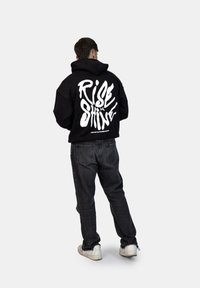 Felpa nera oversize con il testo bianco "RISE & SHINE" sulla schiena. Abbinata a jeans scuri e sneaker bianche. Tessuto morbido, stile casual.