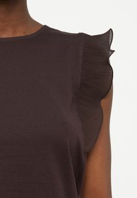 Σκούρο καφέ αμάνικο τοπ με διαφανές, πλισέ βολάν στον ώμο, φορεμένο από άτομο με μέτριου-σκούρου τόνου δέρμα.