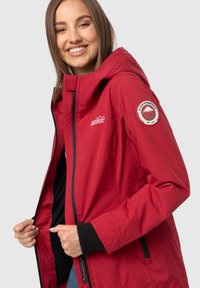 Chaqueta impermeable roja con cierre de cremallera, capucha ajustable y puños de canalé negros. Un parche con el logo en la manga izquierda añade un toque de detalle.
