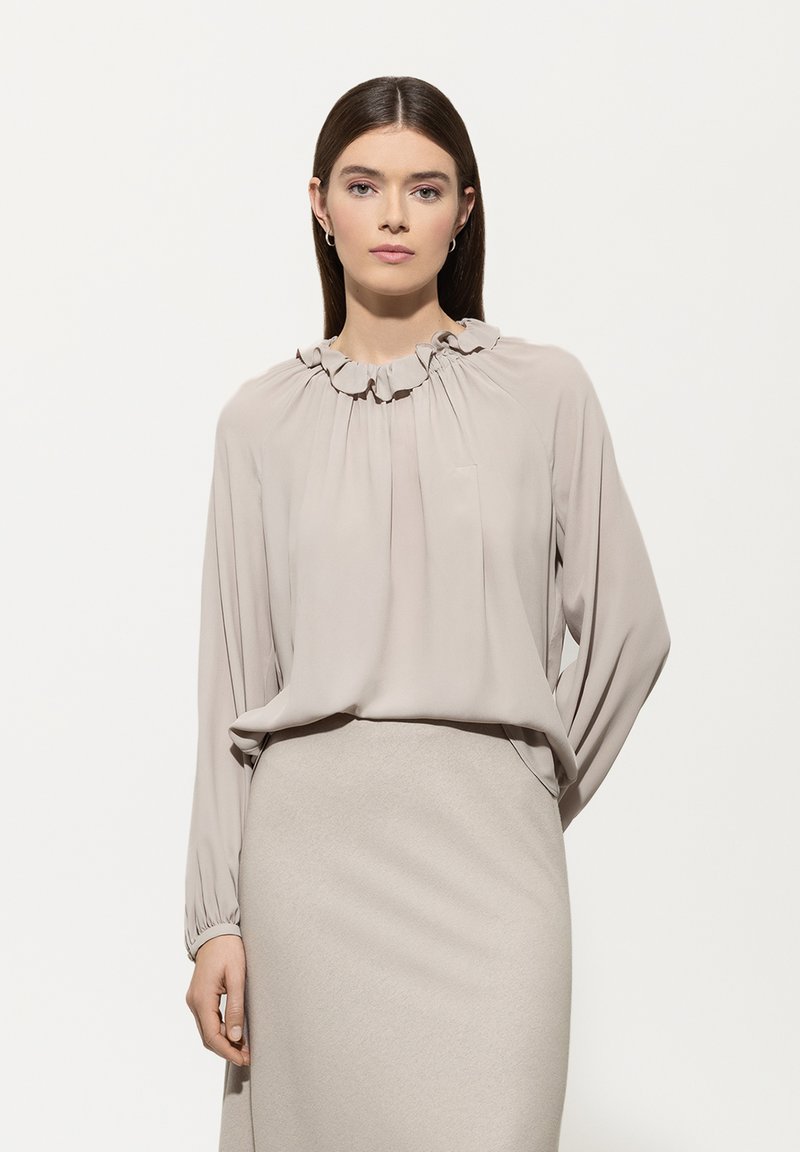 Eine Frau mit glattem braunem Haar trägt eine beige Bluse mit langen Ärmeln und rüschigem Kragen sowie einen passenden Rock und steht vor einem einfarbigen hellen Hintergrund.