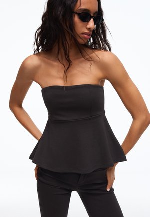 Femeie cu păr lung și închis la culoare, purtând top peplum negru fără bretele, pantaloni negri și ochelari de soare negri, pozând cu mâinile pe șolduri.