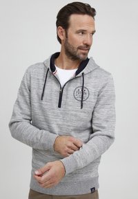 Gray heathered hoodie with a zip neck, navy accents, and a circular logo patch. Features long sleeves and ribbed cuffs. Textured fabric.