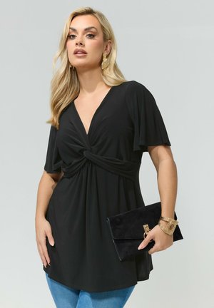 Blonde Frau trägt ein schwarzes, vorne geknotetes Oberteil und blaue Jeans, hält eine schwarze Clutch, accessorisiert mit goldenen Ohrringen und Armband, neutraler Hintergrund.