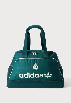 Τουρκουάζ αθλητική τσάντα Adidas με λευκό λογότυπο, έμβλημα της Ρεάλ Μαδρίτης, διπλές χειρολαβές στην κορυφή, θήκες με φερμουάρ και δίχτυα στις πλαϊνές τσέπες.