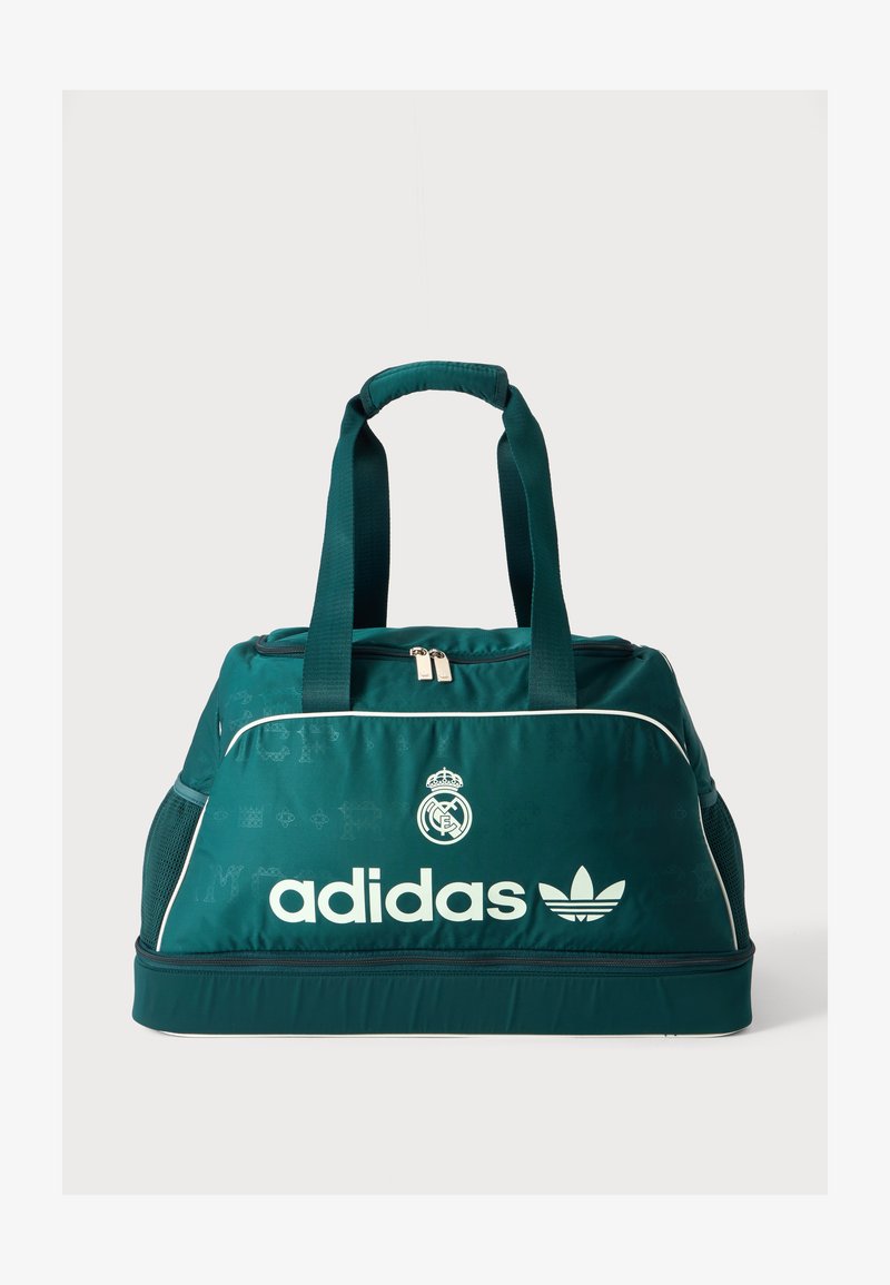 Τουρκουάζ αθλητική τσάντα Adidas με λευκό λογότυπο, έμβλημα της Ρεάλ Μαδρίτης, διπλές χειρολαβές στην κορυφή, θήκες με φερμουάρ και δίχτυα στις πλαϊνές τσέπες.