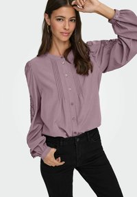 Pika varrukaga lavendli pluus nööpidega ees, volditud detailide ja lillemustriga puff varrukatel, kombineeritud mustade teksadega.