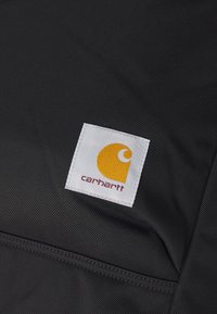 Tissu noir avec une surface texturée, présentant un morceau blanc affichant un logo jaune et "carhartt" en rouge.