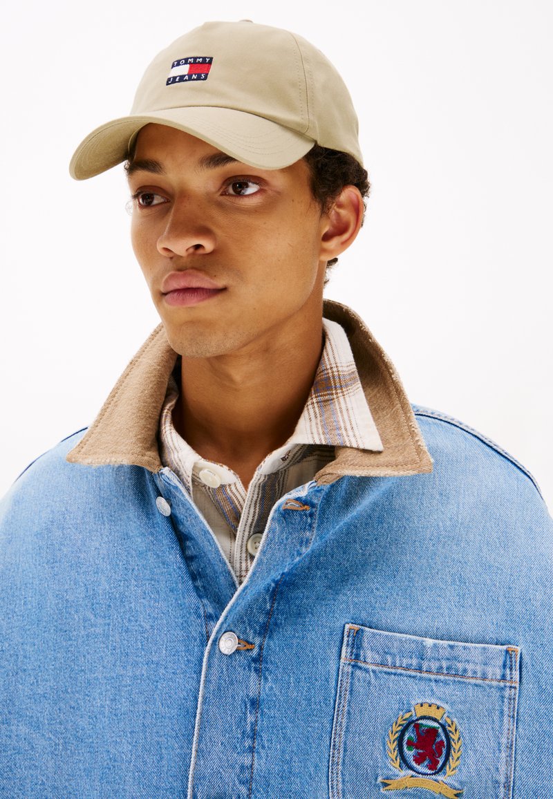 Casquette beige avec le logo "Tommy Jeans", veste en denim avec un col beige, et chemise à carreaux avec un design à boutons. Patch brodé notable sur la veste.