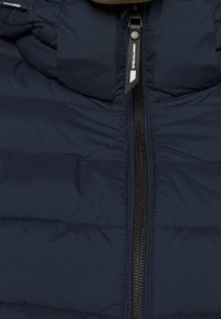 Nahaufnahme einer marineblauen Steppjacke mit zentralem schwarzem Reißverschluss und Zugriegel, der mit "Denim Tom Tailor" beschriftet ist.