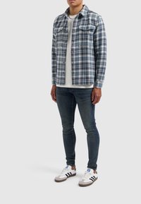 Blauw-witte geruite flanellen shirt met knoopzakken, gedragen over een wit T-shirt, gecombineerd met donkere jeans en witte sneakers met bruine accenten.