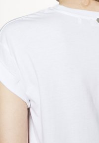 Camiseta blanca de manga corta hecha de una tela suave, con mangas enrolladas y un sutil detalle de botón en la parte posterior del escote.