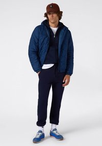 Giacca imbottita blu navy sopra una felpa con cappuccio blu navy, abbinata a pantaloni della tuta blu navy e scarpe da ginnastica blu con dettagli bianchi. Cappello marrone in testa.