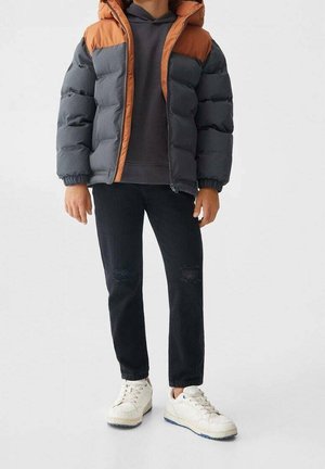 Personne portant une doudoune bicolore orange et grise, un jean foncé déchiré, des baskets blanches et un sweat à capuche foncé sur un fond uni.