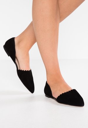 Sorte ruskindsflade sko med spids tå og bølgede udskæringer på siderne, med lav hæl og glat tekstur.