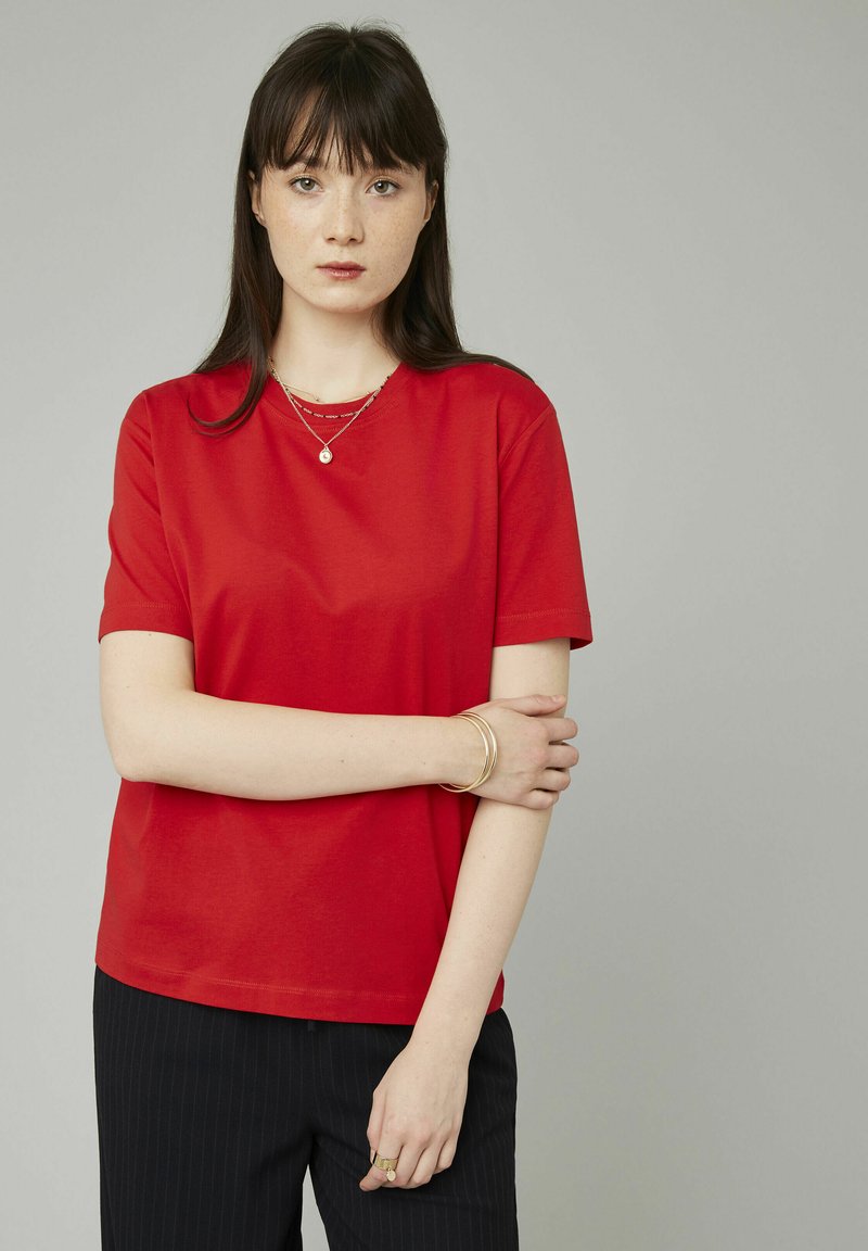 T-shirt en coton rouge à manches courtes et col rond, associé à un pantalon noir à rayures. Les bijoux en or remarquables comprennent des colliers superposés et des bracelets.