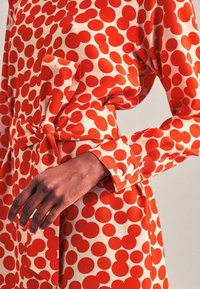 Röd klänning med prickar i sidenliknande tyg. Har ett midjebälte och en knappmanchett på ärmen. Stora, djärva prickar på en krämfärgad bakgrund.