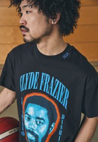 Sort bomulds T-shirt med et grafisk motiv af Clyde Frazier i blå og orange med kontrasterende tekst. Model der holder en basketball.