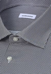 Gros plan d’un col de chemise à motifs avec des boutons blancs et l’étiquette de la marque "seidensticker" cousue à l’intérieur du col.