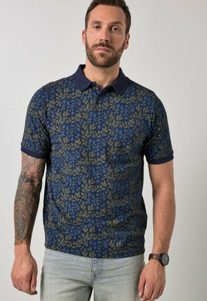 Man met een marineblauw poloshirt met groene en blauwe bladpatronen, lichte spijkerbroek, toont getatoeëerde linker onderarm en een horloge om de rechter pols.