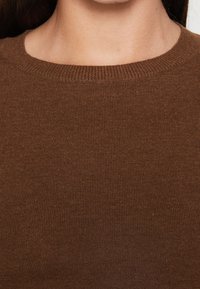 Brązowy sweter z prążkowanego dzianiny z okrągłym dekoltem, prezentujący gładką teksturę i jednolity kolor w całej powierzchni. Brak widocznych wzorów lub elementów metalowych.